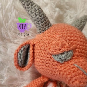 May include: A crocheted plush toy with a gray horn, gray ears, and a peach-colored body. The toy is lying on a white fluffy surface.
