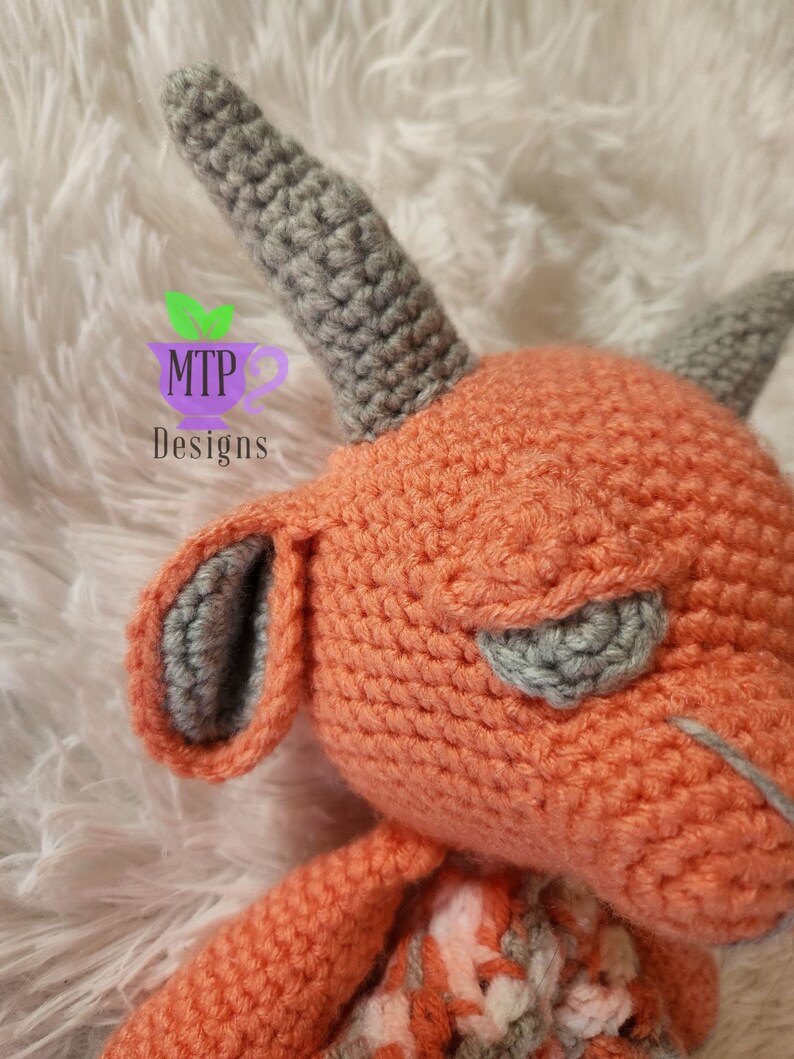 May include: A crocheted plush toy with a gray horn, gray ears, and a peach-colored body. The toy is lying on a white fluffy surface.