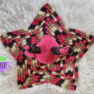 May include: A pink crocheted star-shaped baby blanket with a pink crocheted animal in the center. The blanket has a multi-colored pattern of pink, brown, black, and cream.
