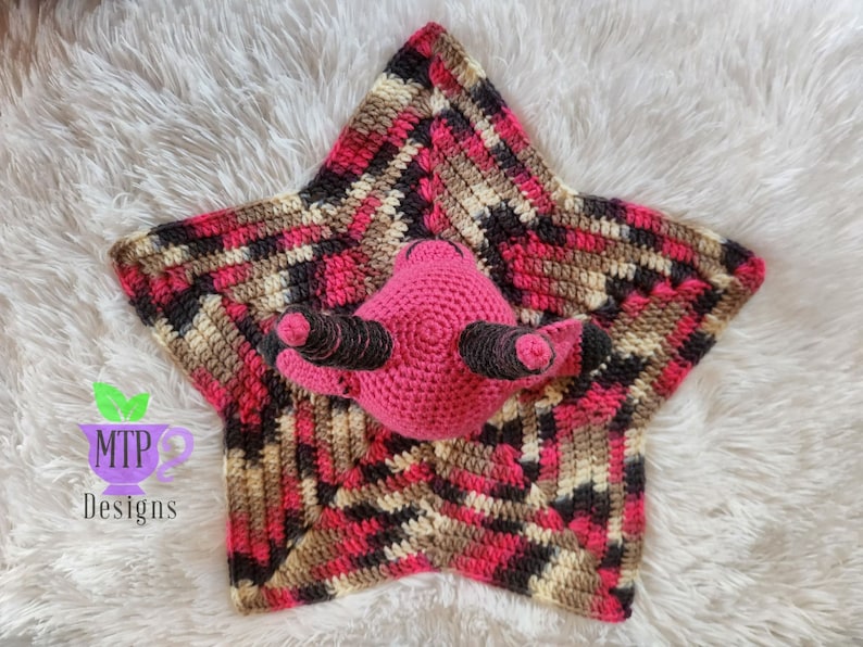 May include: A pink crocheted star-shaped baby blanket with a pink crocheted animal in the center. The blanket has a multi-colored pattern of pink, brown, black, and cream.