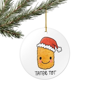 May include: A white, round Christmas ornament featuring a cartoon tater tot wearing a red Santa hat. The tater tot has a smiling face and the words "TATER TOT" are printed below. A gold string is attached for hanging.