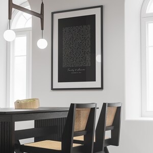 May include: Black and white framed print with a poem about love and a couple's names and wedding date. The print is hanging on a white wall above a black dining table with two black chairs.