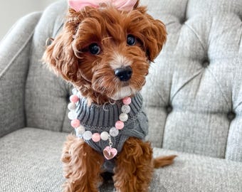 May include: A small, fluffy brown puppy with a pink bow on its head and a gray sweater. The puppy is wearing a necklace with pink and white beads and a heart pendant. It is sitting on a gray upholstered chair.