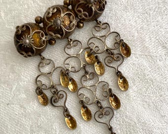 May include: An antique brooch featuring three ornate, filigree spheres with intricate detailing. Below, a silver-toned metal design with dangling gold-toned teardrop-shaped accents. The brooch has a vintage aesthetic.