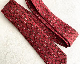 May include: A red necktie featuring a repeating pattern of dark brown rope-like chains and thin gold-colored vertical lines arranged in a diamond grid. The tie is folded over itself, displaying the pointed end and the fabric texture clearly.