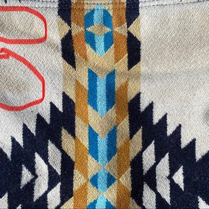 May include: Close-up of a woven textile with a geometric pattern. The design features repeating shapes in navy, white, tan, brown, blue, and gold. The fabric has a textured appearance, with some discoloration visible.
