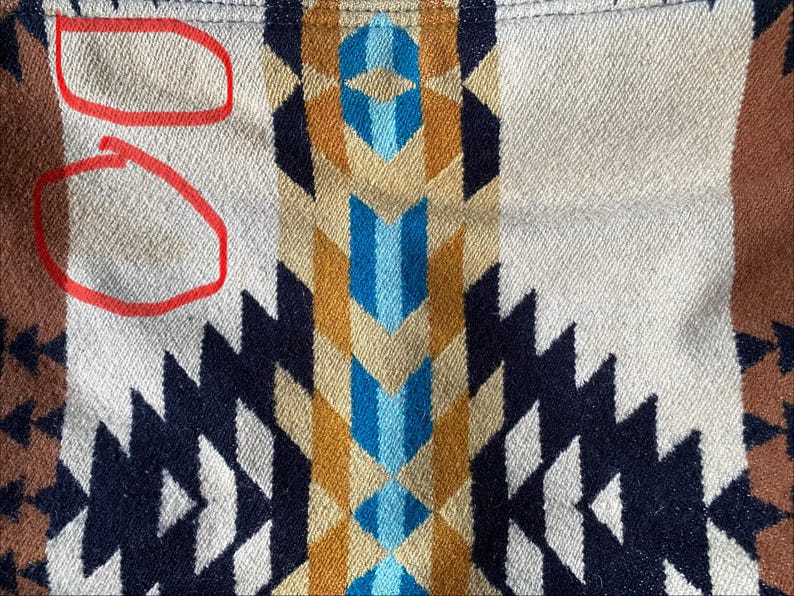 May include: Close-up of a woven textile with a geometric pattern. The design features repeating shapes in navy, white, tan, brown, blue, and gold. The fabric has a textured appearance, with some discoloration visible.
