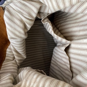 May include: Close-up of the interior of a brown suede bag. The inside is lined with a cream-colored fabric with thin, vertical brown stripes. A pocket is visible on the side. The bag's opening is visible at the top.