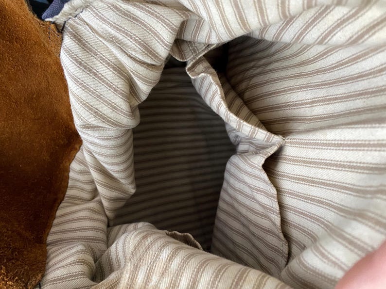 May include: Close-up of the interior of a brown suede bag. The inside is lined with a cream-colored fabric with thin, vertical brown stripes. A pocket is visible on the side. The bag's opening is visible at the top.