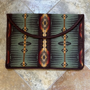 Peut inclure: Une pochette rectangulaire en tissu avec un motif du Sud-Ouest américain dans des tons de vert, marron et rouge. Elle est bordée d'une garniture marron foncé et se ferme par un rabat. Le motif comprend des éléments géométriques et floraux stylisés.