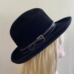 Puede incluir: Sombrero fedora negro con una banda de cuero y hebilla. El sombrero tiene una copa redondeada y un ala ancha. La banda de cuero está cosida con hilo blanco.