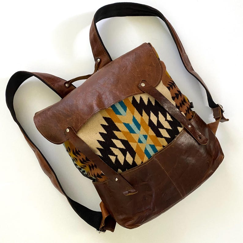 May include: A brown leather backpack with a flap closure. The front panel features a geometric pattern in beige, black, yellow, and teal. The bag has adjustable black straps and brown leather accents.