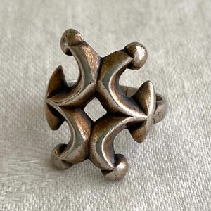 Puede incluir: Un anillo plateado con un diseño ornamentado y simétrico. El anillo presenta un patrón repetitivo de formas curvas y acentos redondeados, creando una estética geométrica audaz. El metal tiene una apariencia ligeramente envejecida, lo que aumenta su atractivo vintage.