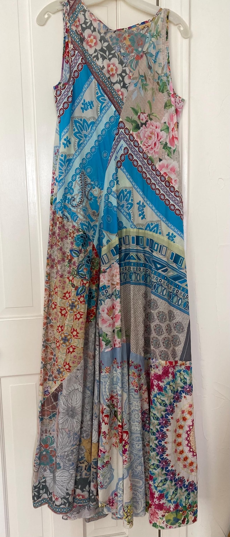 Puede incluir: Un vestido largo sin mangas con un dise&ntilde;o de patchwork. El vestido presenta una variedad de estampados, incluyendo estampados florales, paisley y geom&eacute;tricos en tonos de azul, rosa y beige. El vestido est&aacute; colgado en una percha blanca.