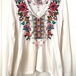 May include: White long-sleeve shirt with a V-neck and colorful floral embroidery. The embroidery features red, pink, blue, and green flowers with intricate details. The shirt has a relaxed fit and a slightly asymmetrical hemline.