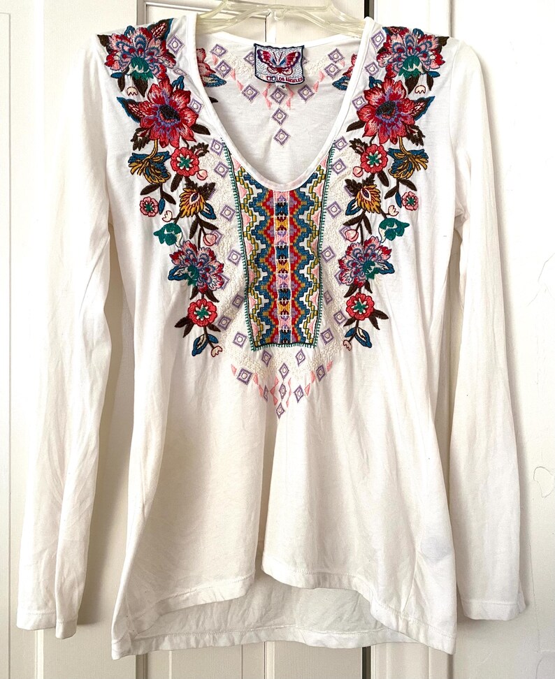 May include: White long-sleeve shirt with a V-neck and colorful floral embroidery. The embroidery features red, pink, blue, and green flowers with intricate details. The shirt has a relaxed fit and a slightly asymmetrical hemline.