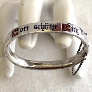 Puede incluir: Una pulsera articulada de plata con la frase alemana "Gott schütze Dich" en letras azules. La pulsera tiene detalles de esmalte rojo y una delicada cadena.