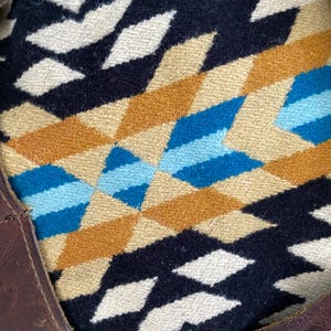 May include: A brown leather tote bag with a geometric patterned textile interior. The textile features a repeating diamond design in shades of black, tan, blue, and white. The bag's leather has visible stitching and metal rivets.