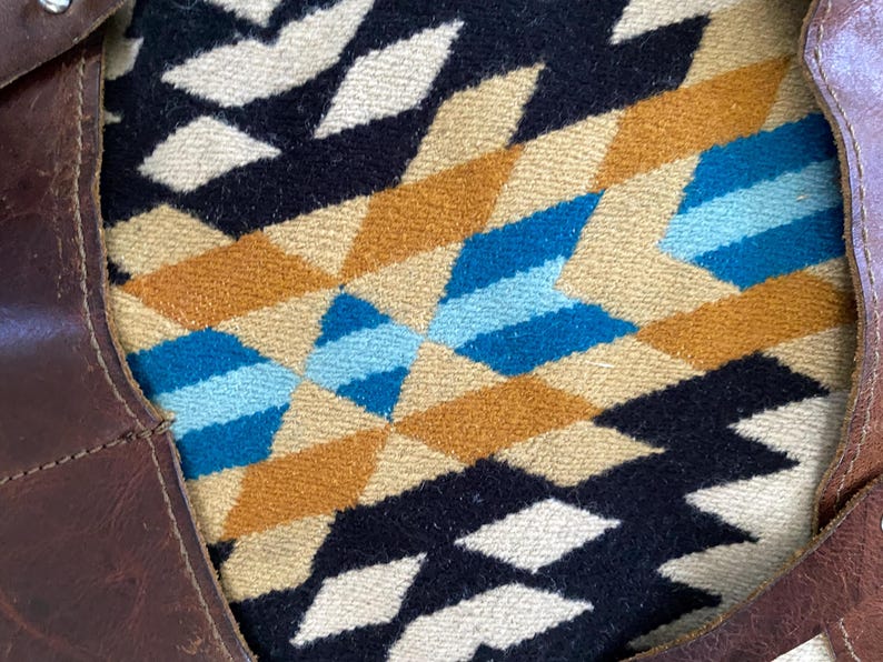 May include: A brown leather tote bag with a geometric patterned textile interior. The textile features a repeating diamond design in shades of black, tan, blue, and white. The bag's leather has visible stitching and metal rivets.