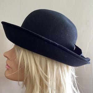 Puede incluir: Un sombrero de fieltro negro con ala ancha y corona baja. El sombrero tiene una banda de terciopelo negro alrededor del ala.