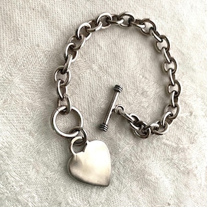 Puede incluir: Un brazalete de plata con un dije de corazón. El brazalete presenta un diseño de eslabones de cadena y un cierre de palanca. El dije de corazón es liso y pulido. El brazalete probablemente está hecho de plata de ley.
