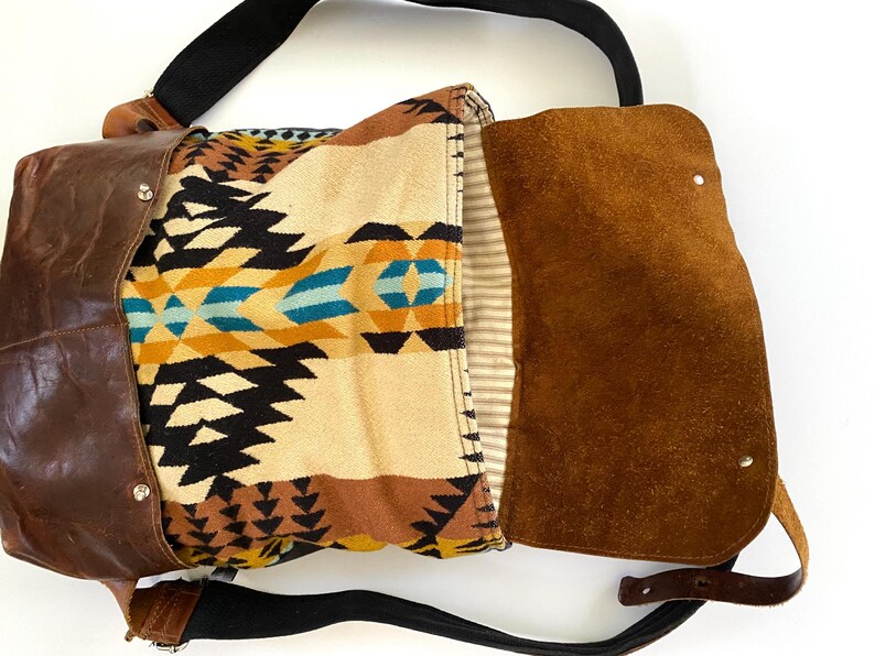 May include: A brown leather messenger bag with a flap closure. The bag features a patterned fabric interior with black, yellow, and turquoise geometric designs. It has a black shoulder strap and a brown leather exterior.