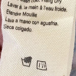Puede incluir: Primer plano de una etiqueta de tela blanca con instrucciones de cuidado en ingl&eacute;s, franc&eacute;s y espa&ntilde;ol. El texto dice "Lava a mano con agua fr&iacute;a. Seca colgado". Incluye s&iacute;mbolos de lavado.