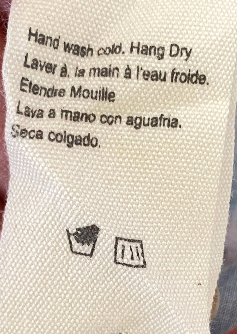 Puede incluir: Primer plano de una etiqueta de tela blanca con instrucciones de cuidado en ingl&eacute;s, franc&eacute;s y espa&ntilde;ol. El texto dice "Lava a mano con agua fr&iacute;a. Seca colgado". Incluye s&iacute;mbolos de lavado.