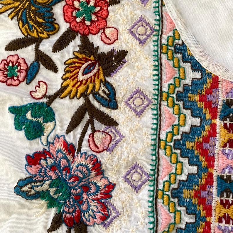May include: Close-up of a white garment with colorful floral embroidery. The embroidery features red, pink, yellow, and blue flowers with brown stems and green leaves. The design includes geometric patterns and a decorative border.