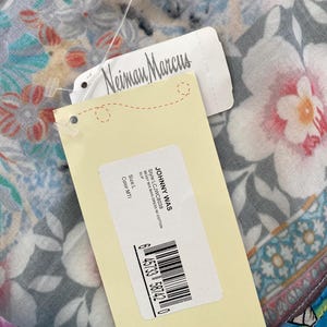 Puede incluir: Un vestido largo con estampado floral con etiquetas de Johnny Was y Neiman Marcus. El vestido presenta un colorido estampado floral. La etiqueta indica el estilo LCJWC8038, talla L y color MTI. El sitio web www.johnnywas.com es visible.