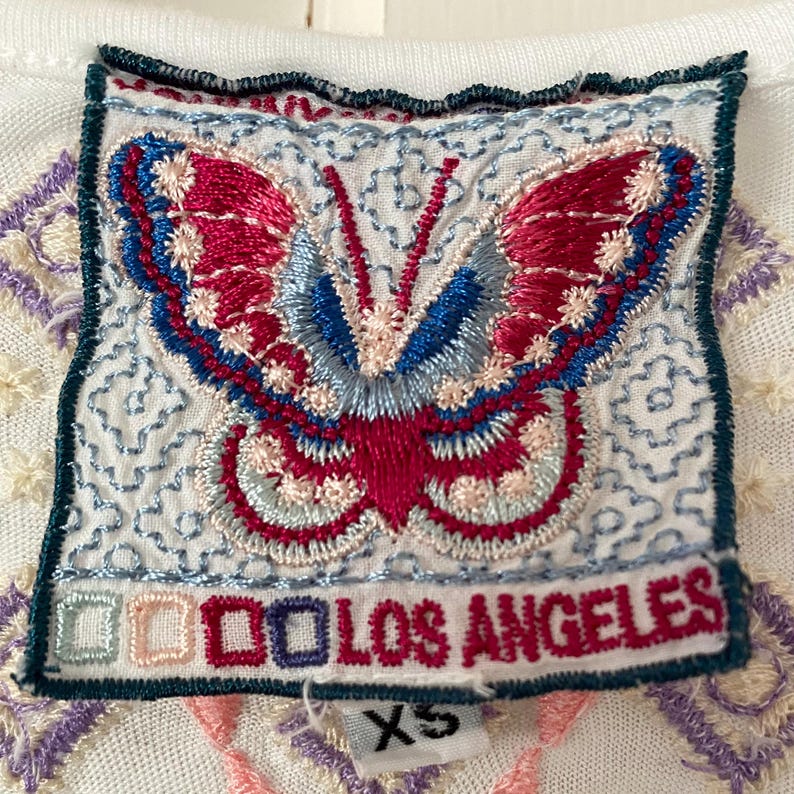 May include: Close-up of a white top with an embroidered patch featuring a butterfly design. The butterfly has red, blue, and pink details, with the text "LOS ANGELES" below. The patch is bordered with a dark green trim, and the shirt is size XS.