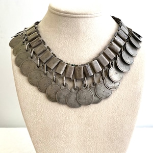 May include: A silver necklace with multiple rows of round, coin-shaped pendants. The necklace is made of metal and has a vintage look.