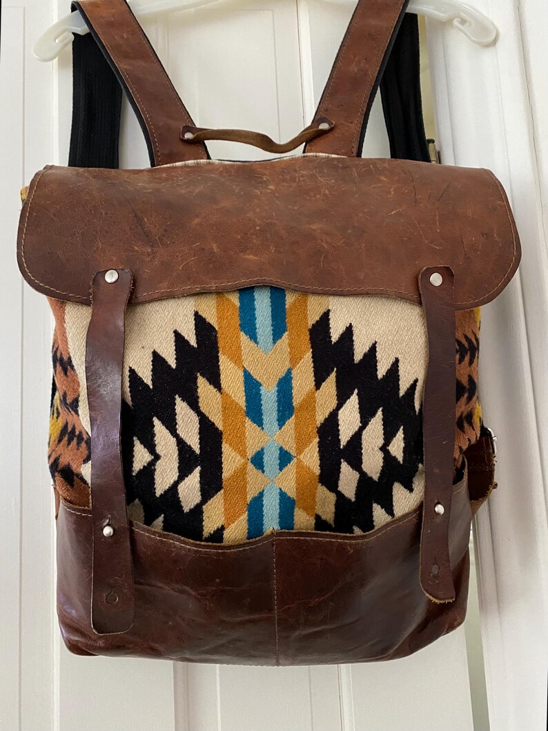 May include: A brown leather backpack with a geometric patterned fabric center. The fabric features black, white, yellow, and blue diamond shapes. The bag has brown leather straps and a flap closure. The backpack is hanging on a white door.