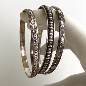 May include: Three silver-toned metal bangles featuring distinct textures. One bangle has an intricate floral vine pattern, another displays a repeating horizontal ribbed design, and the third features a series of embossed heart and flower motifs.