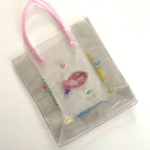 May include: Clear tote bag with pink handles and decorative fish and floral designs. The bag features colorful accents and text in a cursive font. The bag is made of a transparent material.