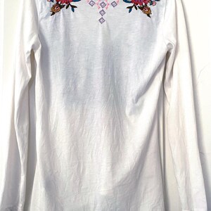 May include: White long-sleeve shirt with floral embroidery in shades of red, pink, blue, and green on the shoulders. The embroidery includes a geometric design in pink and purple. The shirt has a scoop neck.