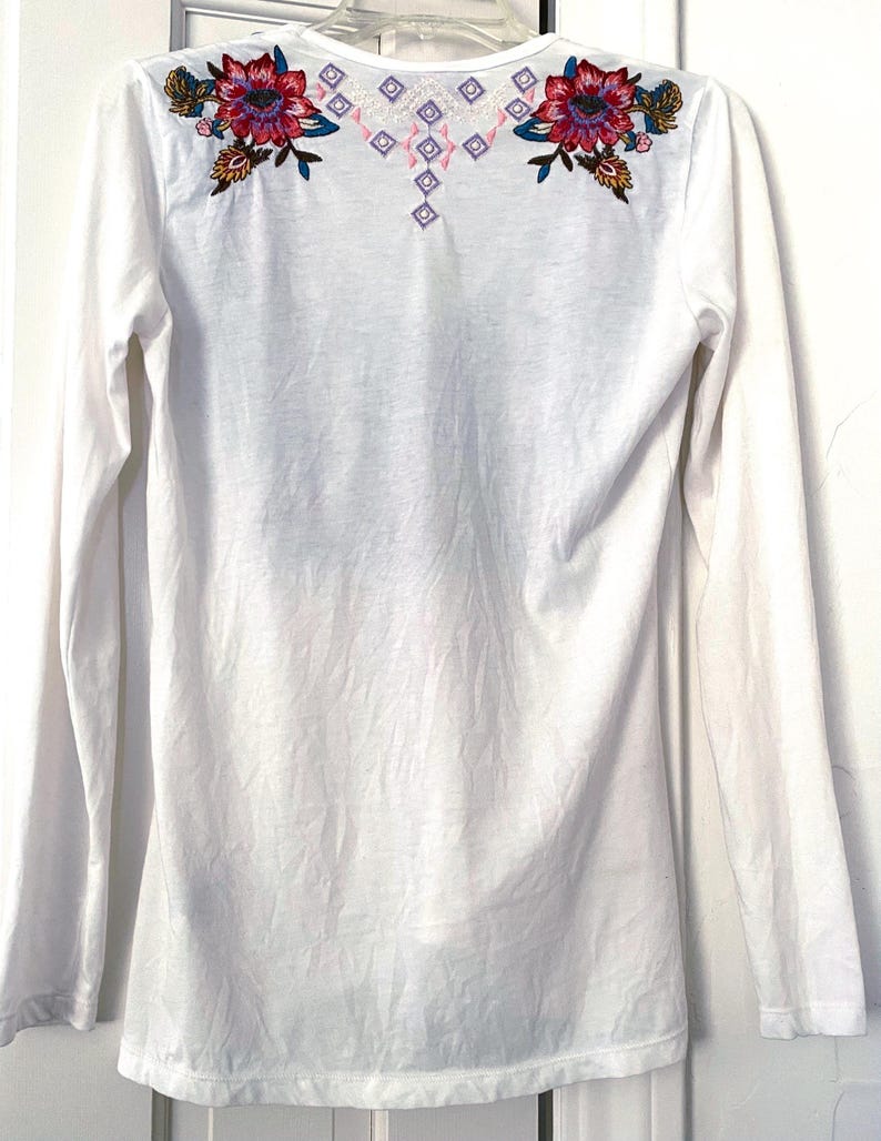 May include: White long-sleeve shirt with floral embroidery in shades of red, pink, blue, and green on the shoulders. The embroidery includes a geometric design in pink and purple. The shirt has a scoop neck.