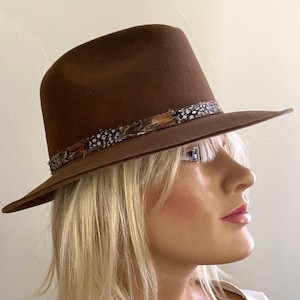 May include: A brown felt fedora hat featuring a structured crown and a wide brim. The base of the crown is accented with a decorative band composed of small, multi-coloured feathers with brown, black, and white speckled patterns throughout.