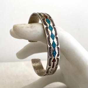 May include: A silver cuff bracelet featuring a central row of diamond-shaped turquoise stone inlays and two red stone inlays at the ends. The metal surface is decorated with stamped feather-like patterns along the edges of the band.