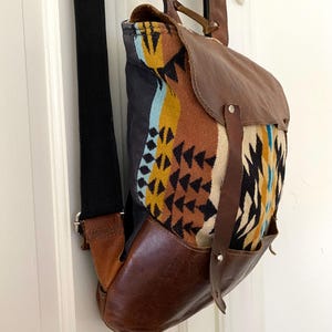 May include: A brown leather backpack with a flap closure and a patterned fabric front. The fabric features a geometric design in shades of yellow, blue, and brown. The bag has a black strap and leather accents.