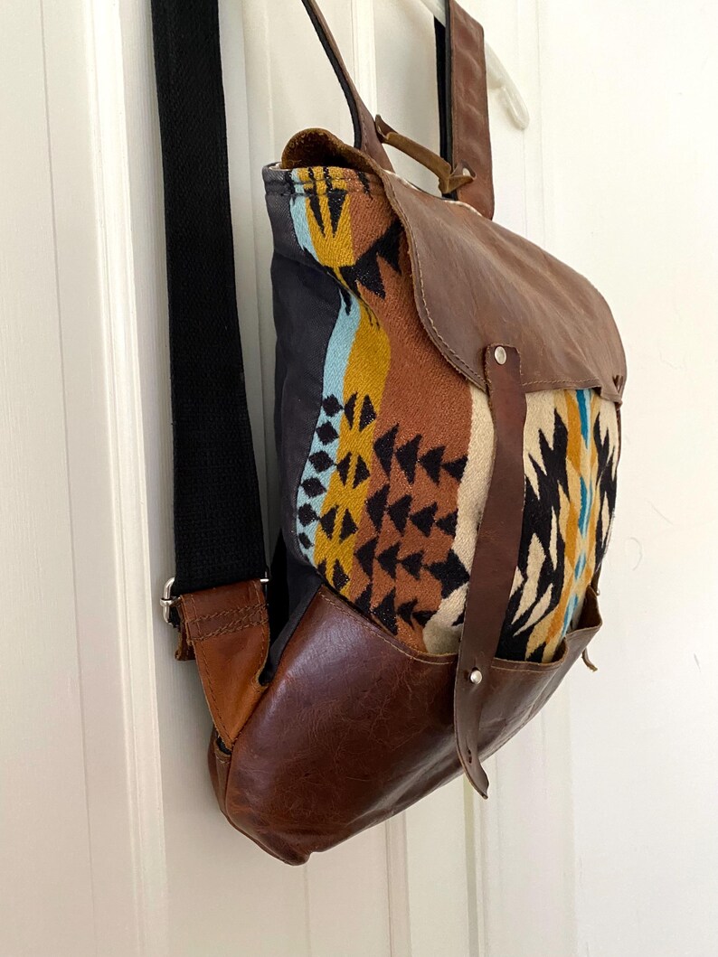 May include: A brown leather backpack with a flap closure and a patterned fabric front. The fabric features a geometric design in shades of yellow, blue, and brown. The bag has a black strap and leather accents.