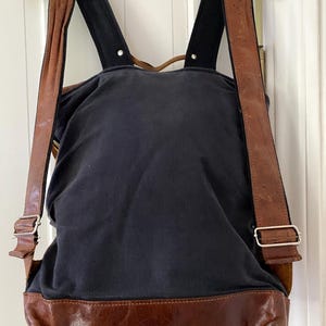 May include: A navy blue backpack with brown leather accents. The bottom and straps are brown leather, and the straps are adjustable with silver buckles. The backpack is hanging on a white hanger.