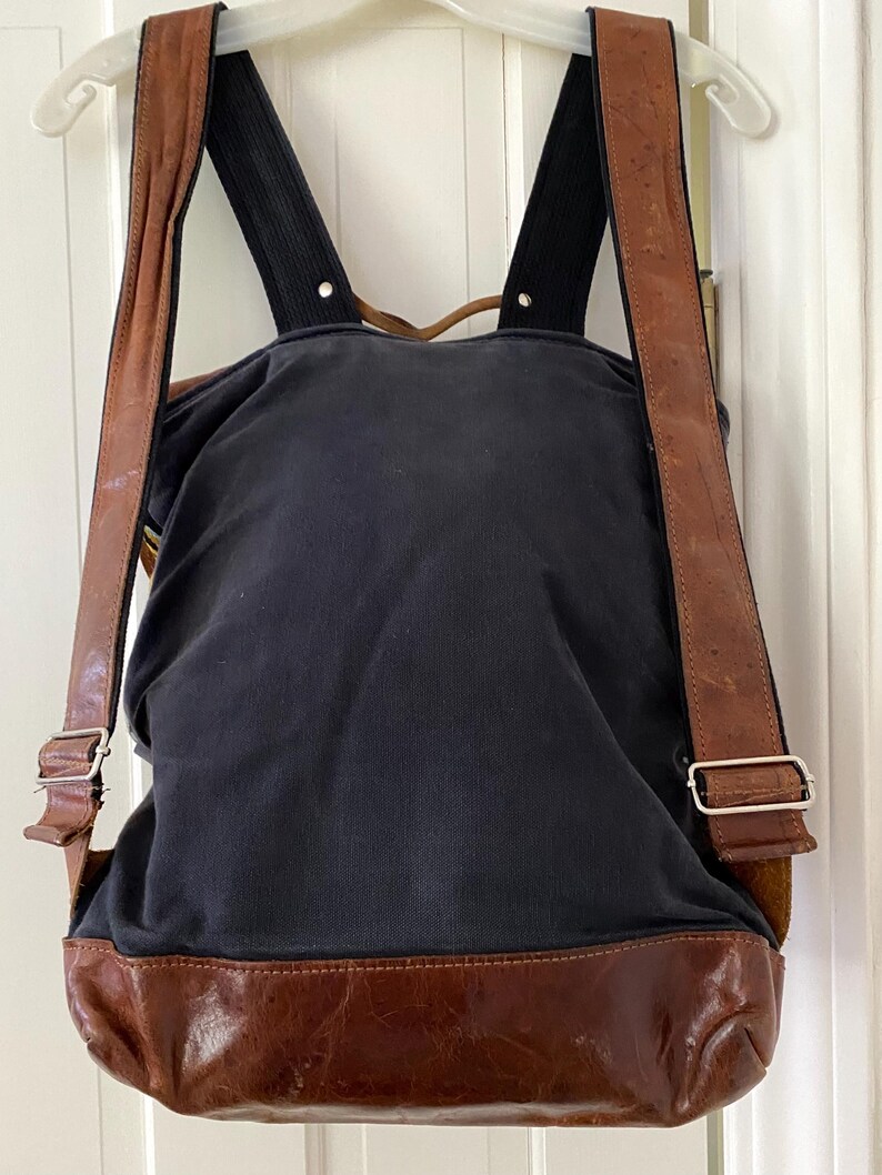 May include: A navy blue backpack with brown leather accents. The bottom and straps are brown leather, and the straps are adjustable with silver buckles. The backpack is hanging on a white hanger.