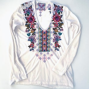 May include: White long-sleeve shirt with a deep V-neck and colorful floral embroidery. The embroidery features pink, purple, blue, and green flowers and geometric patterns. The shirt has a relaxed fit and is made of a soft material.