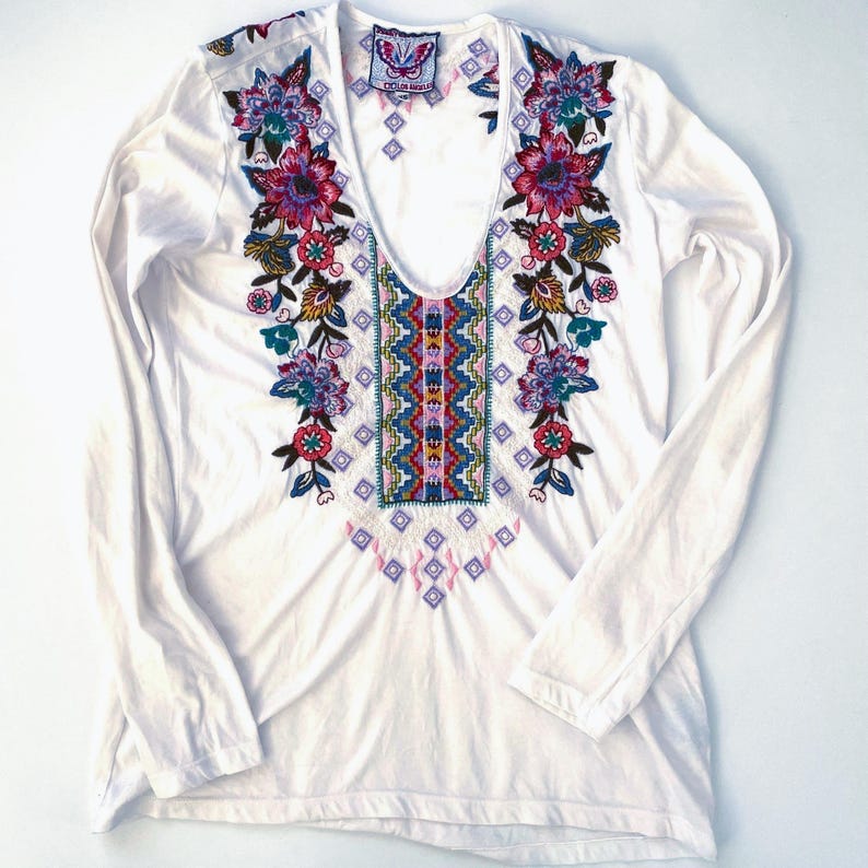 May include: White long-sleeve shirt with a deep V-neck and colorful floral embroidery. The embroidery features pink, purple, blue, and green flowers and geometric patterns. The shirt has a relaxed fit and is made of a soft material.