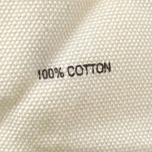 Puede incluir: Primer plano de una tela color crema con el texto "100% COTTON" impreso en negro. La tela tiene una apariencia texturizada y tejida, lo que sugiere un textil de fibra natural.