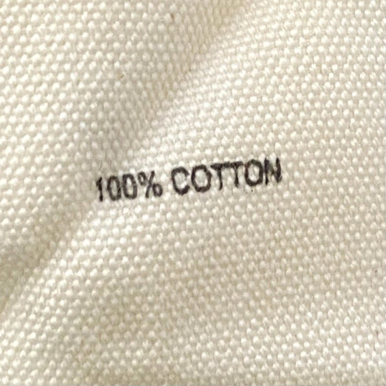 Puede incluir: Primer plano de una tela color crema con el texto "100% COTTON" impreso en negro. La tela tiene una apariencia texturizada y tejida, lo que sugiere un textil de fibra natural.