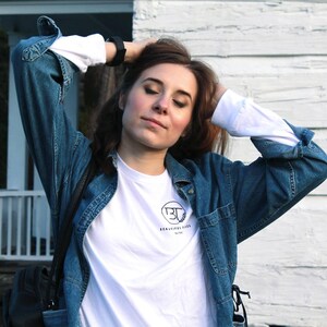 May include: A woman wearing a white t-shirt with a black logo that says "Beautiful Things by BT" and a blue denim jacket.