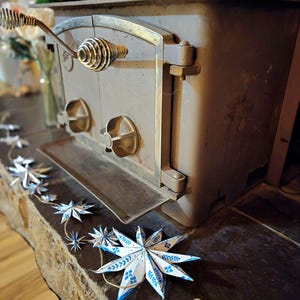 May include: A vintage wood-burning stove with a metal door and a decorative handle. A string of blue and white paper stars hangs in front of the stove.