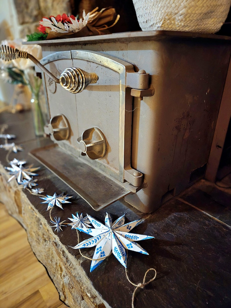 May include: A vintage wood-burning stove with a metal door and a decorative handle. A string of blue and white paper stars hangs in front of the stove.